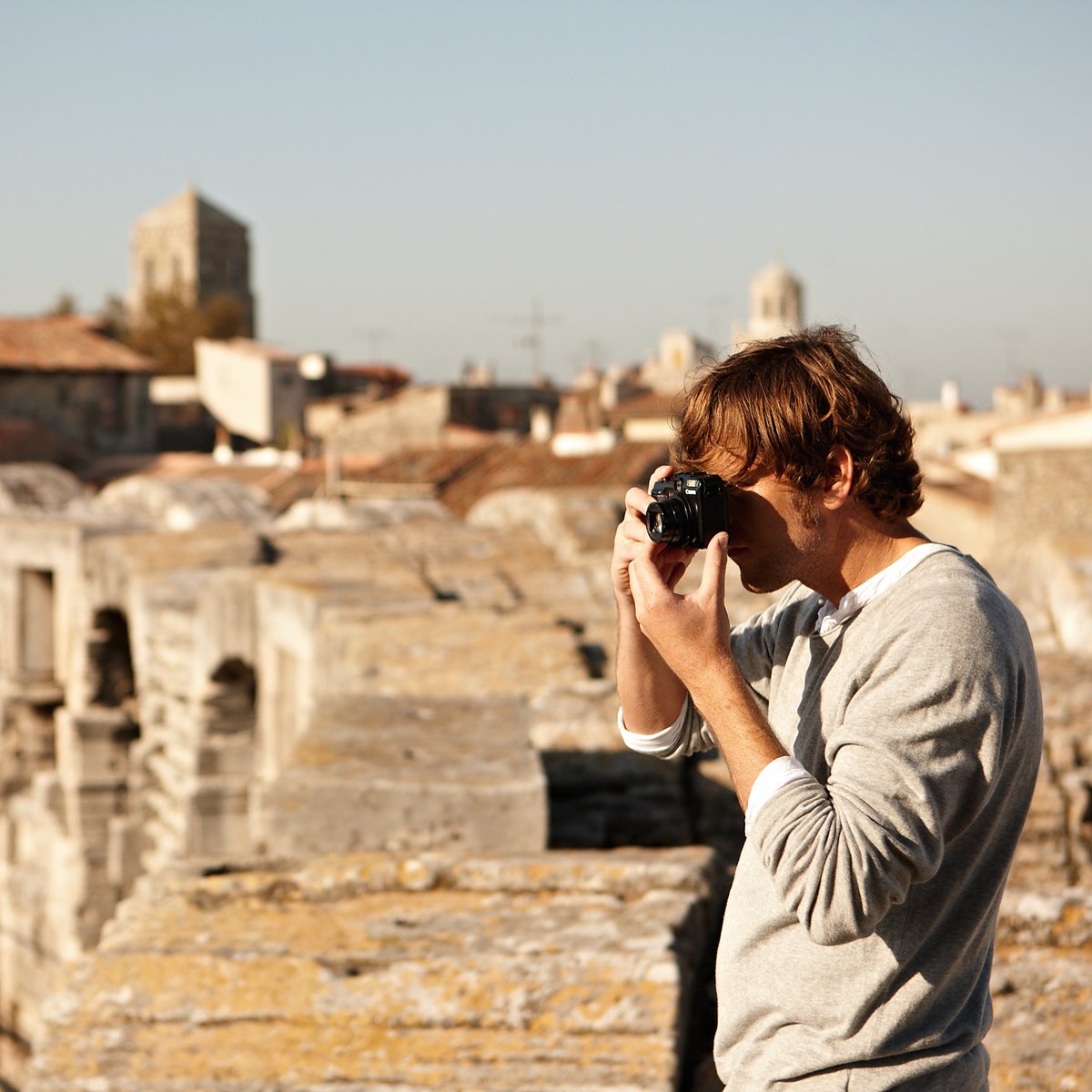 Ein Mann steht auf einer historischen Mauer in Arles und fotografiert etwas. Im Hintergrund die gelben Gebäude der Stadt.