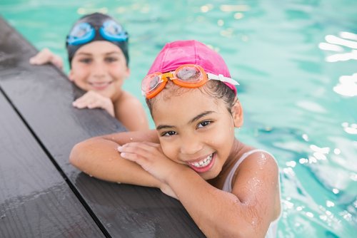 Zwei Kinder mit Badekappen und Schwimmbrillen ruhen sich am Beckenrand eines Pools aus und lächeln ind die Kamera
