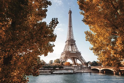 Eiffelturm in Paris im Herbst, umgeben von Bäumen mit orangefarbenen Blättern, Fluss und Brücke im Vordergrund.